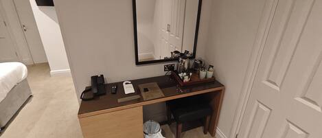 A compact hotel room with a wooden desk, a mirror, a telephone, a trash bin, and a bed.