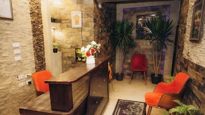 Un área de recepción con un mostrador de madera, dos sillones naranja, una planta en maceta y un tapete decorativo.