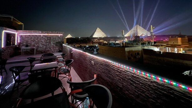 Terraza en la azotea con mesas y sillas, vista a las pirámides y una guirnalda de luces de colores.