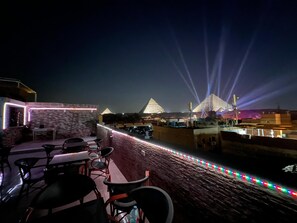 A rooftop terrace with tables and chairs, a view of the pyramids, and a string of colorful lights. - Pyramids Live View Hotel (Giza)