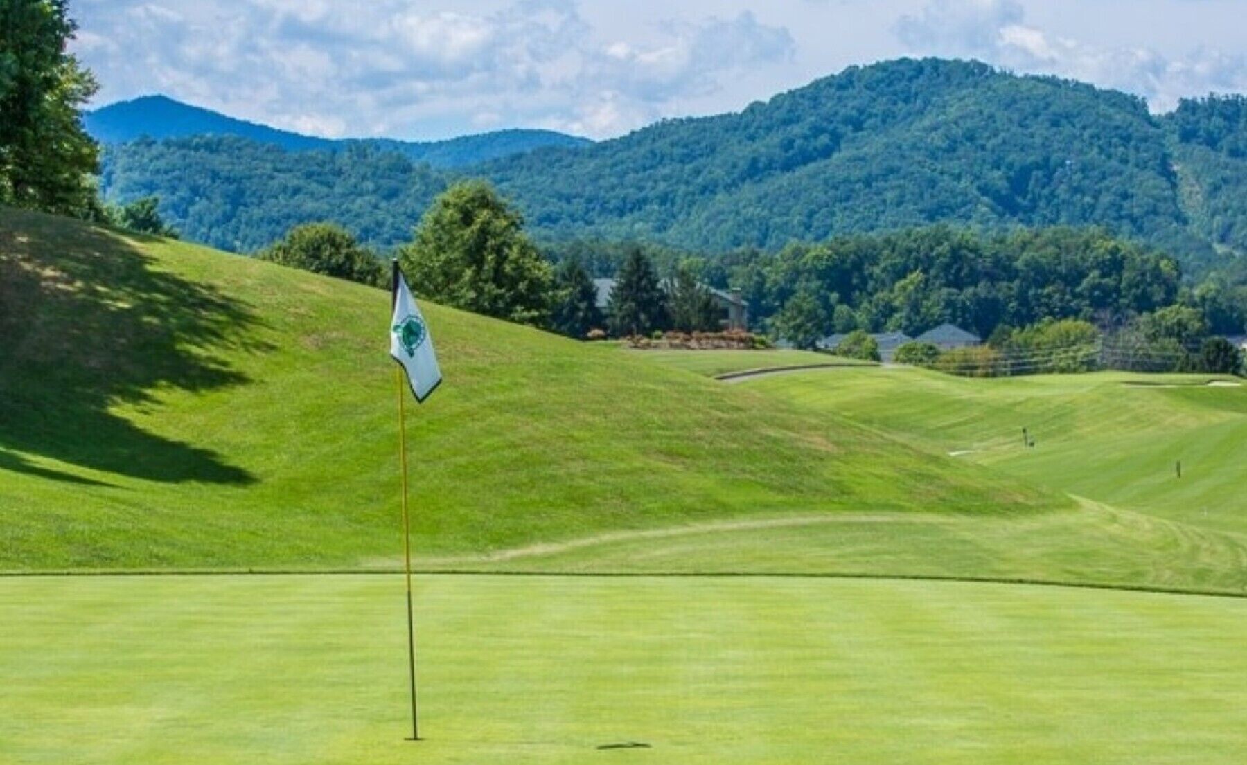 A golf course with a flag on a flagstick, surrounded by rolling green hills and a forested mountain in the background.