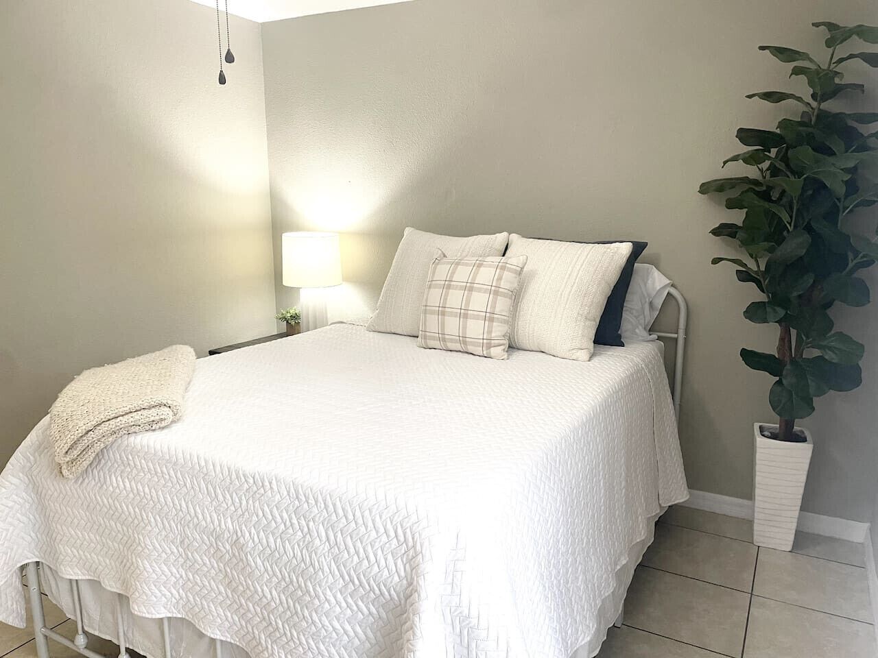 A neatly made bed with a white quilt and pillows, a bedside lamp, and a potted plant beside the bed.