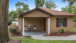 A single-story house with a brown roof and beige walls, number 801, featuring a covered porch with two chairs and a small table, surrounded by greenery and trees.