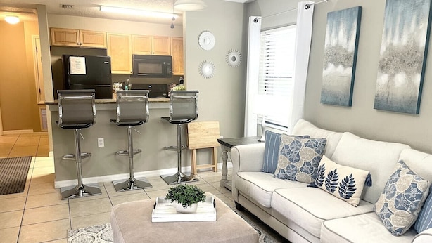 A modern living room with a sofa, two bar stools, a coffee table with a plant, and a kitchen in the background.