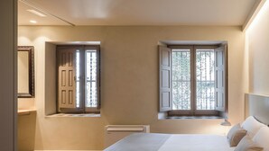 Une chambre à coucher avec un lit, deux fenêtres avec des volets, un radiateur et un miroir au mur.