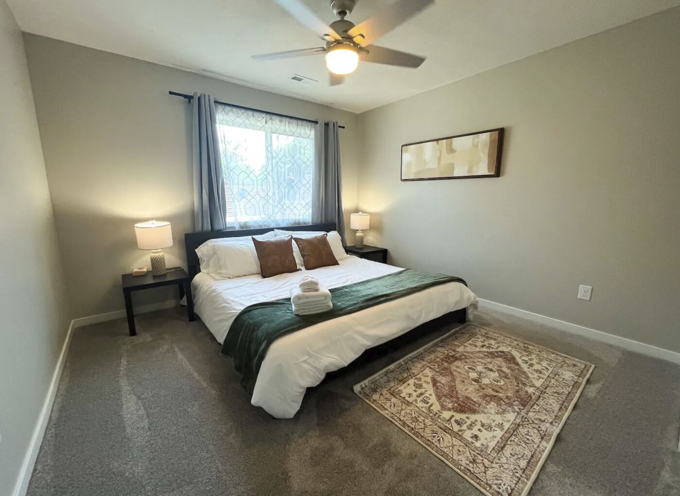 Une chambre à coucher avec un lit, deux tables de chevet avec des lampes, un ventilateur de plafond, une fenêtre avec des rideaux, un tapis et un tableau encadré au mur.
