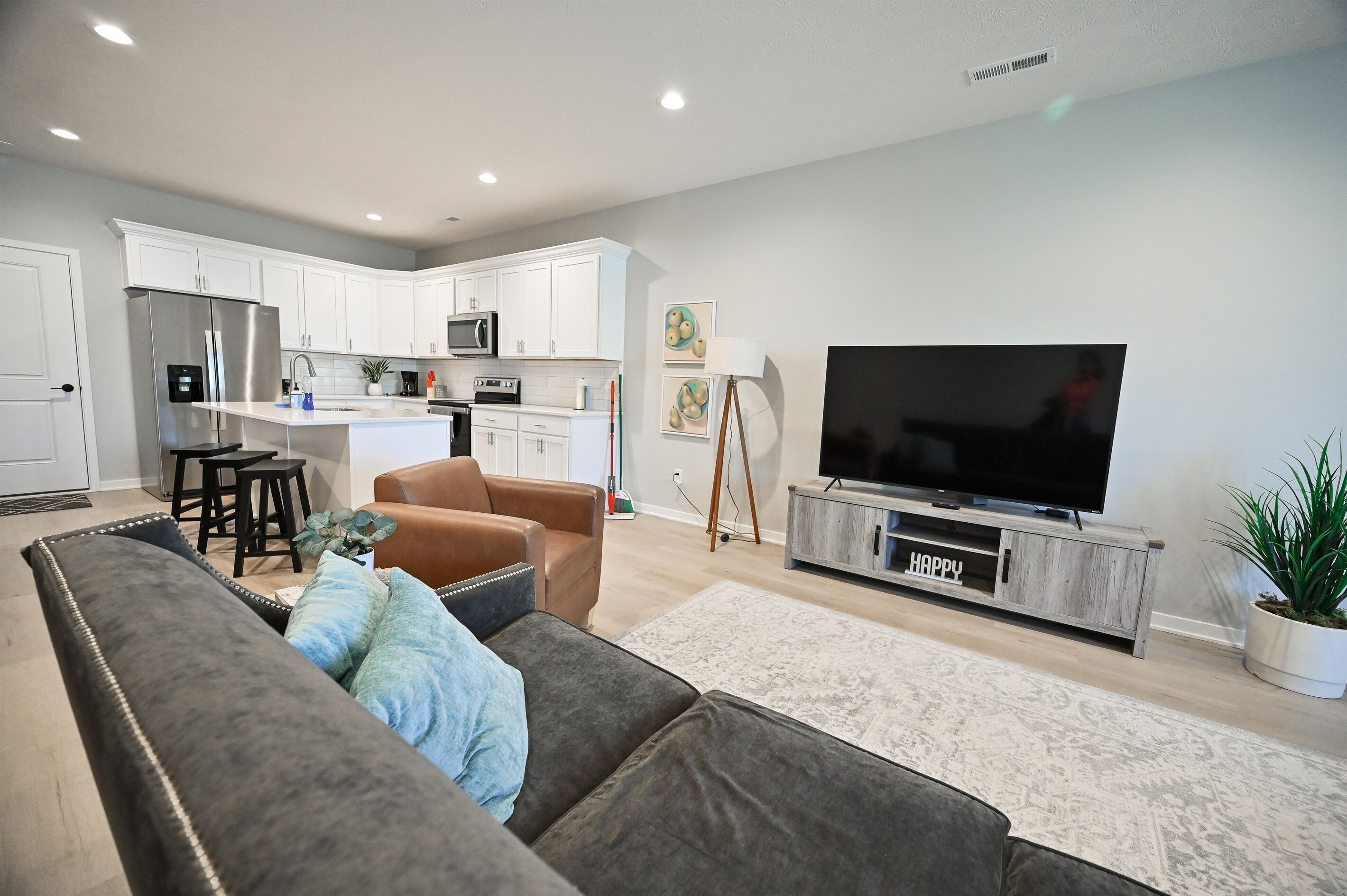 A modern kitchen with white cabinets, a stainless steel refrigerator, and a microwave. There is a dining area with a small table and chairs. A large flat-screen TV is mounted on a wooden cabinet.