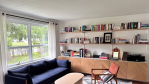 A modern living room with a dark blue sofa, a wooden coffee table, and a red armchair. There is a bookshelf filled with books and a small table with a lamp and a framed picture.