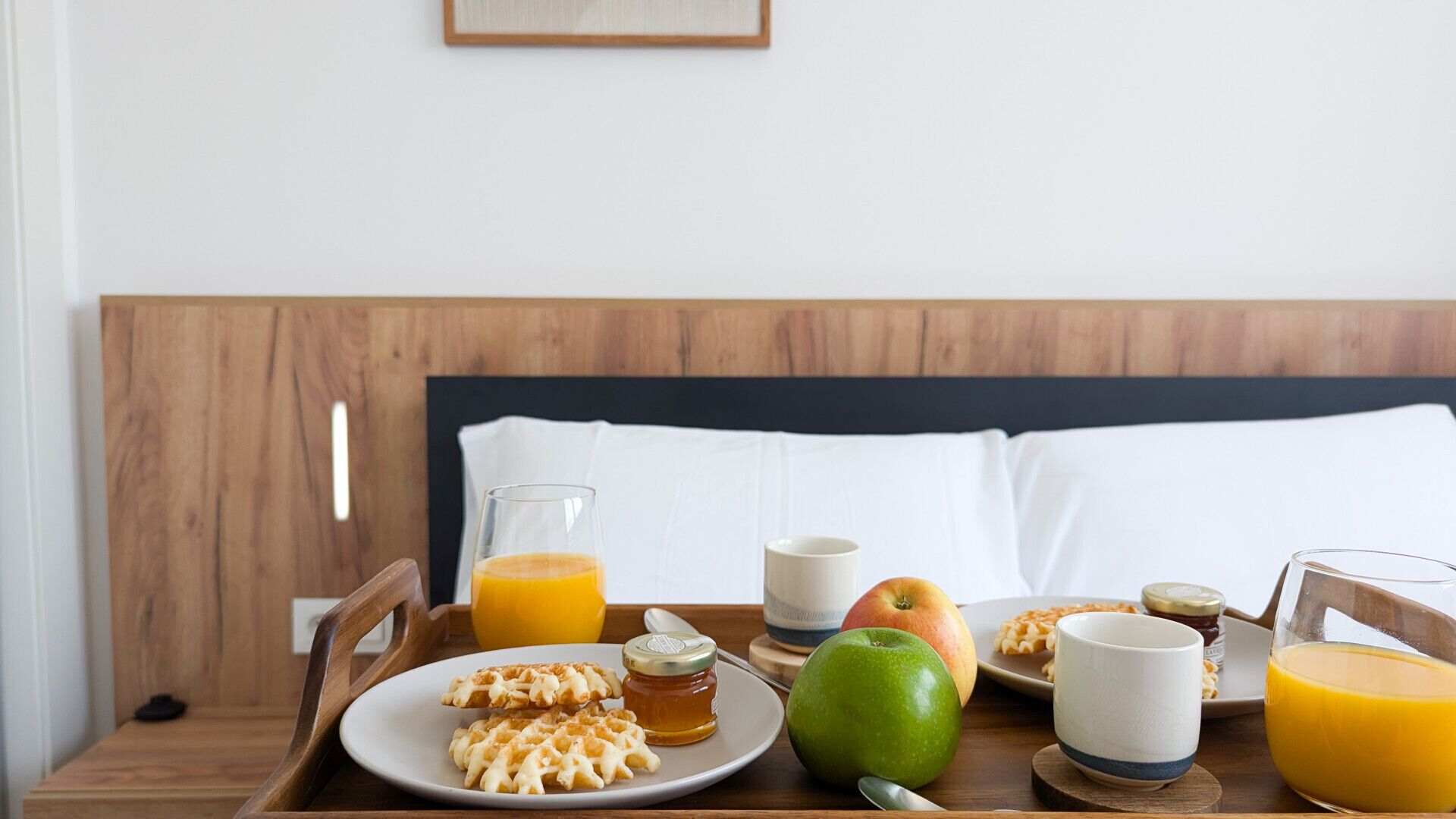A breakfast tray with waffles, apples, honey, and orange juice on a wooden table in a hotel room.