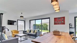 A modern living room with a sofa, coffee table, and dining table. There are large windows, a TV, and a painting on the wall.
