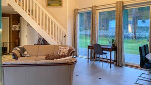 En stue med sofa, spiseplads med bord og stole, trappe og store vinduer med gardiner.