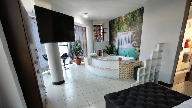 A hotel room with a large bed, a sitting area with a sofa, a small table, a potted plant, a bathtub, and a mural of a waterfall. - Hotel Platinium Palace (Cusco)