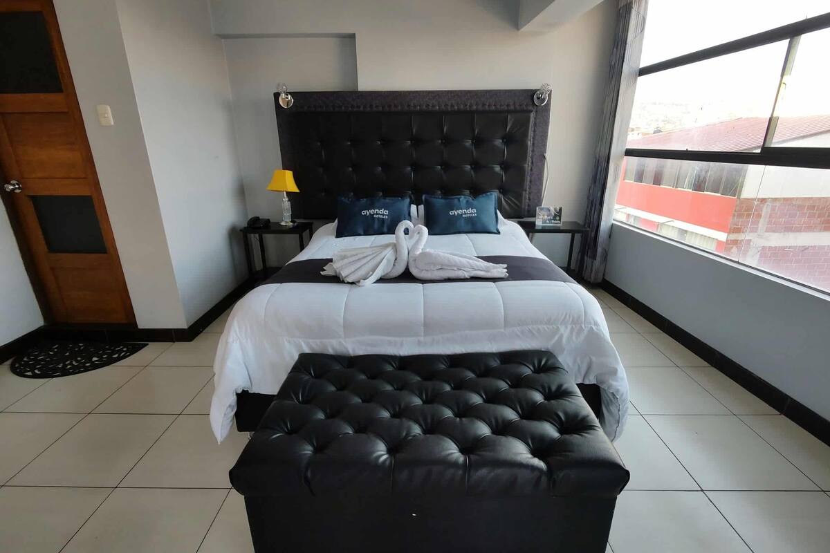 A hotel room with a large bed, a black upholstered bench, a nightstand with a lamp, and a view of the city through the window.
