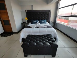 A hotel room with a large bed, a black upholstered bench, a nightstand with a lamp, and a view of the city through the window. - Hotel Platinium Palace (Cusco)
