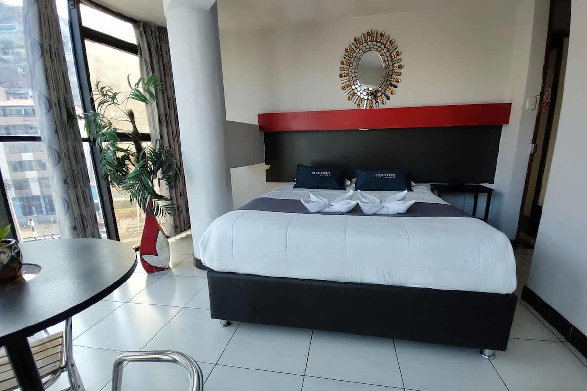 A hotel room with a bed, a round mirror, a red accent wall, and a view through a window.