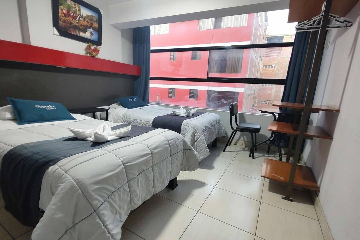 A hotel room with two beds, a desk, and a chair. There is a window with a view of buildings outside.