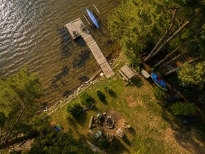 An aerial view of a lakeside property with a wooden dock, a small boat, and a fire pit area.