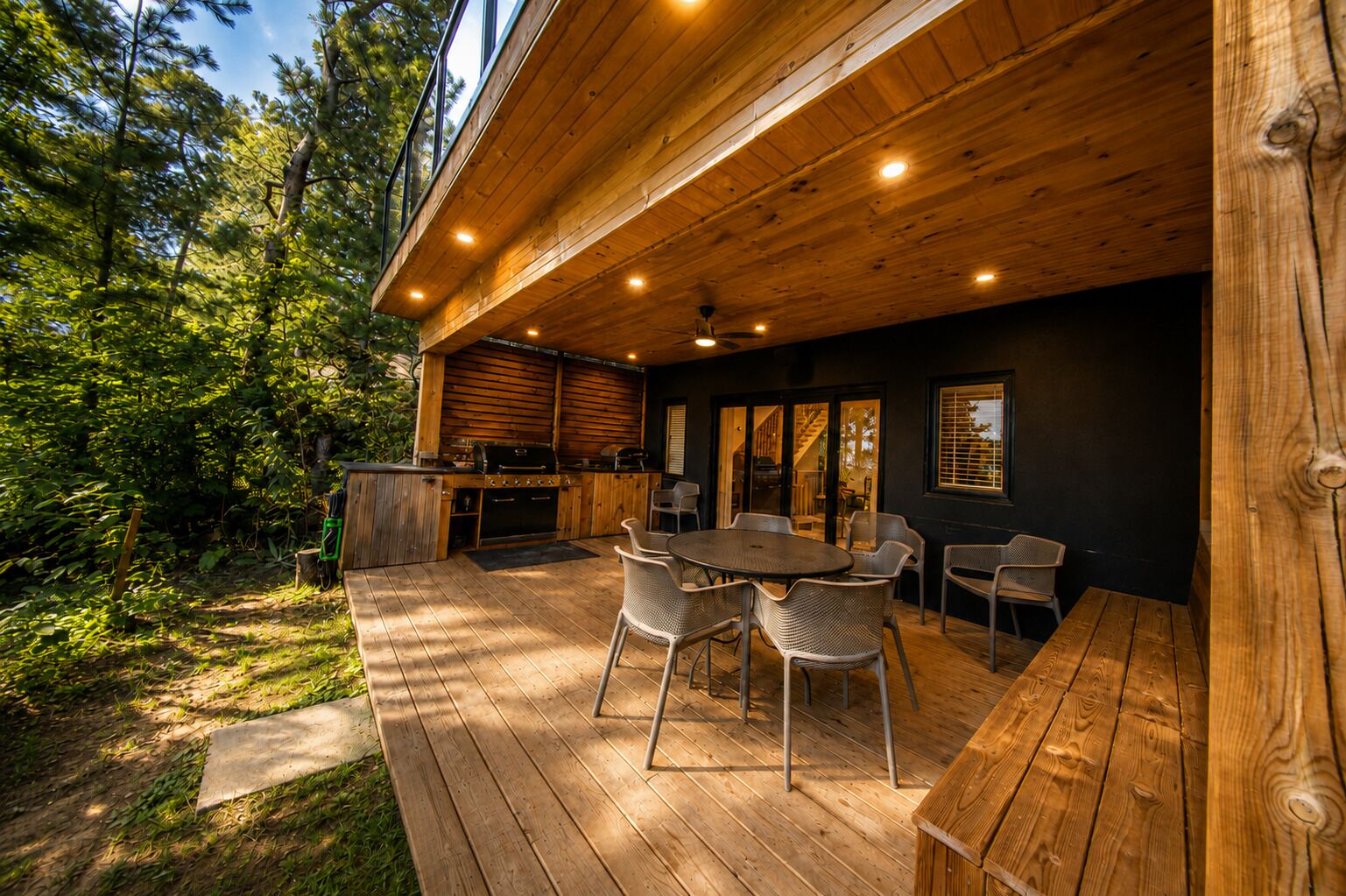 Een terras buiten met een houten tafel en stoelen, een barbecue en uitzicht op het omliggende bos.