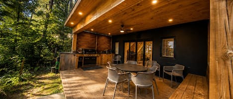 Een terras buiten met een houten tafel en stoelen, een barbecue en uitzicht op het omliggende bos.