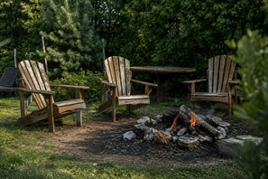 Una fogata con leña, rodeada de sillas Adirondack y una mesa de madera.
