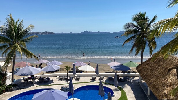 A pool area with multiple pools, umbrellas, and lounge chairs, overlooking a beach with palm trees and a clear blue sky. - Breathtaking ocean front condominium in the gated community of Club Santiago (Manzanillo)