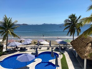A pool area with multiple pools, umbrellas, and lounge chairs, overlooking a beach with palm trees and a clear blue sky. - Stunning apartment with ocean views in the gated Club Santiago community (Manzanillo)