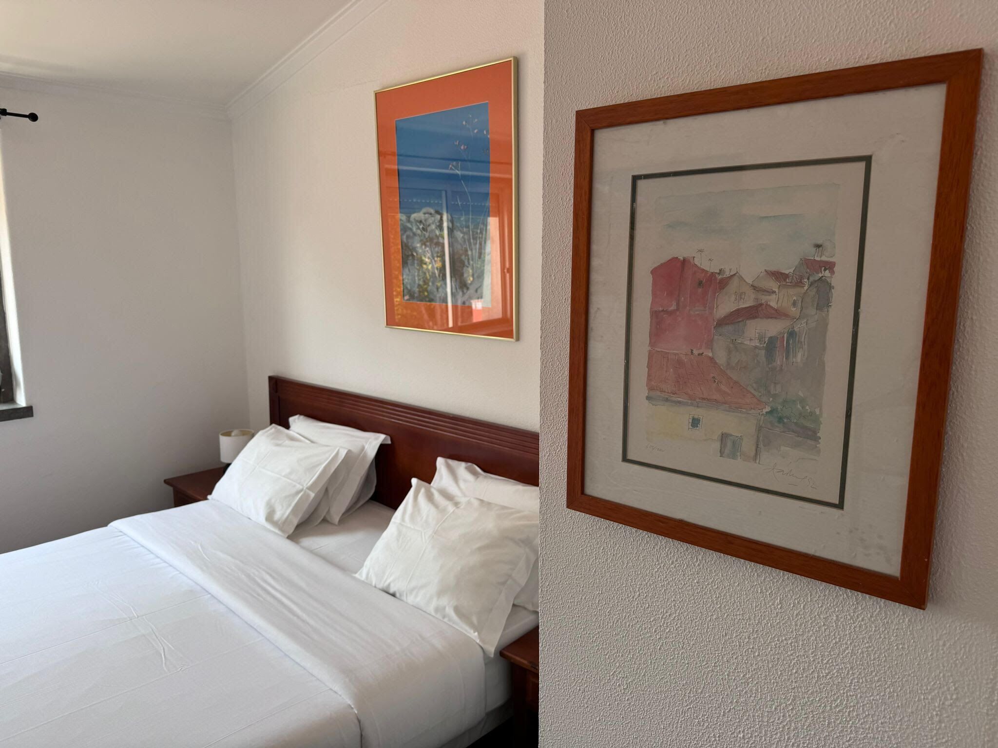 A neatly made bed with white linens, a wooden headboard, and two framed artworks on the wall.