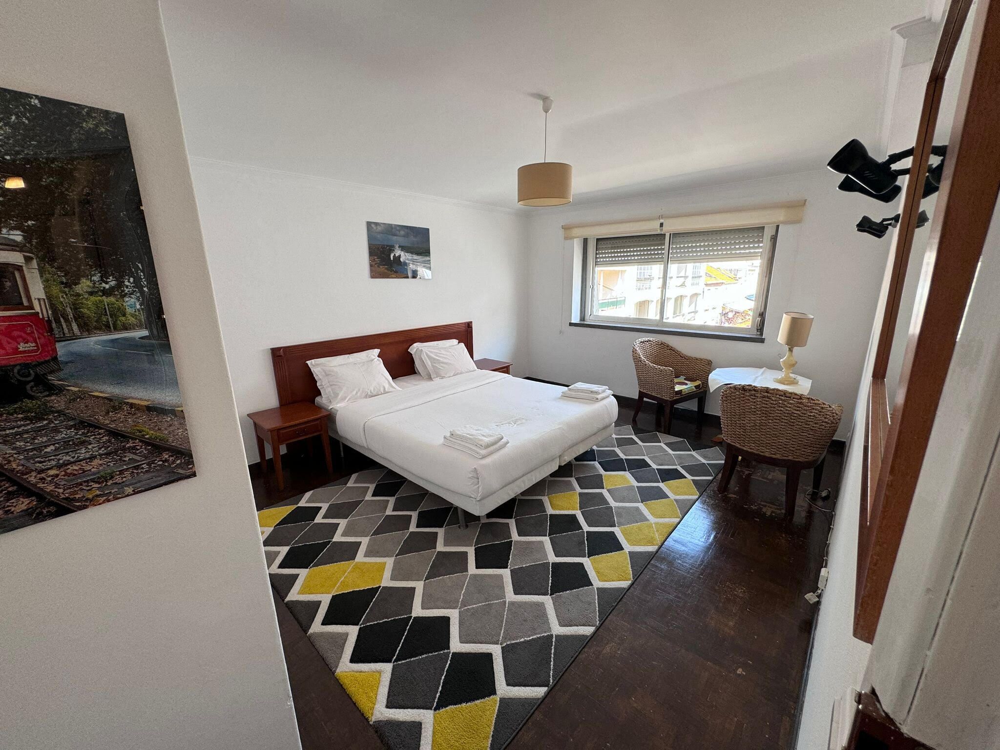A hotel room with a bed, a chair, a table, a window with blinds, and a painting on the wall.