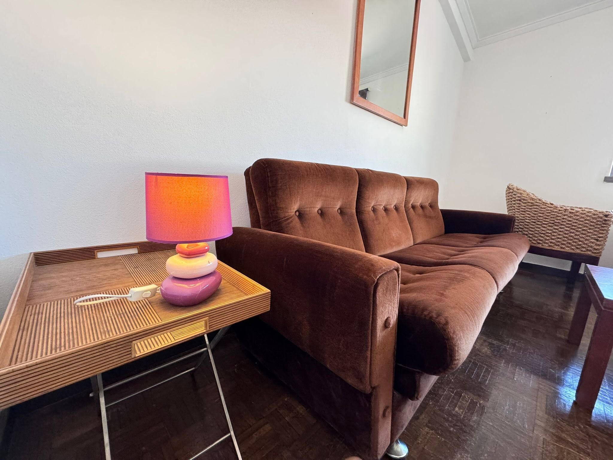 A brown sofa, a wooden side table with a lamp, and a woven chair in a room with wooden flooring and a white wall.