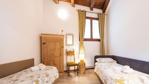 Une chambre avec deux lits simples, une armoire en bois, une petite table avec une lampe et une fenêtre avec des rideaux.