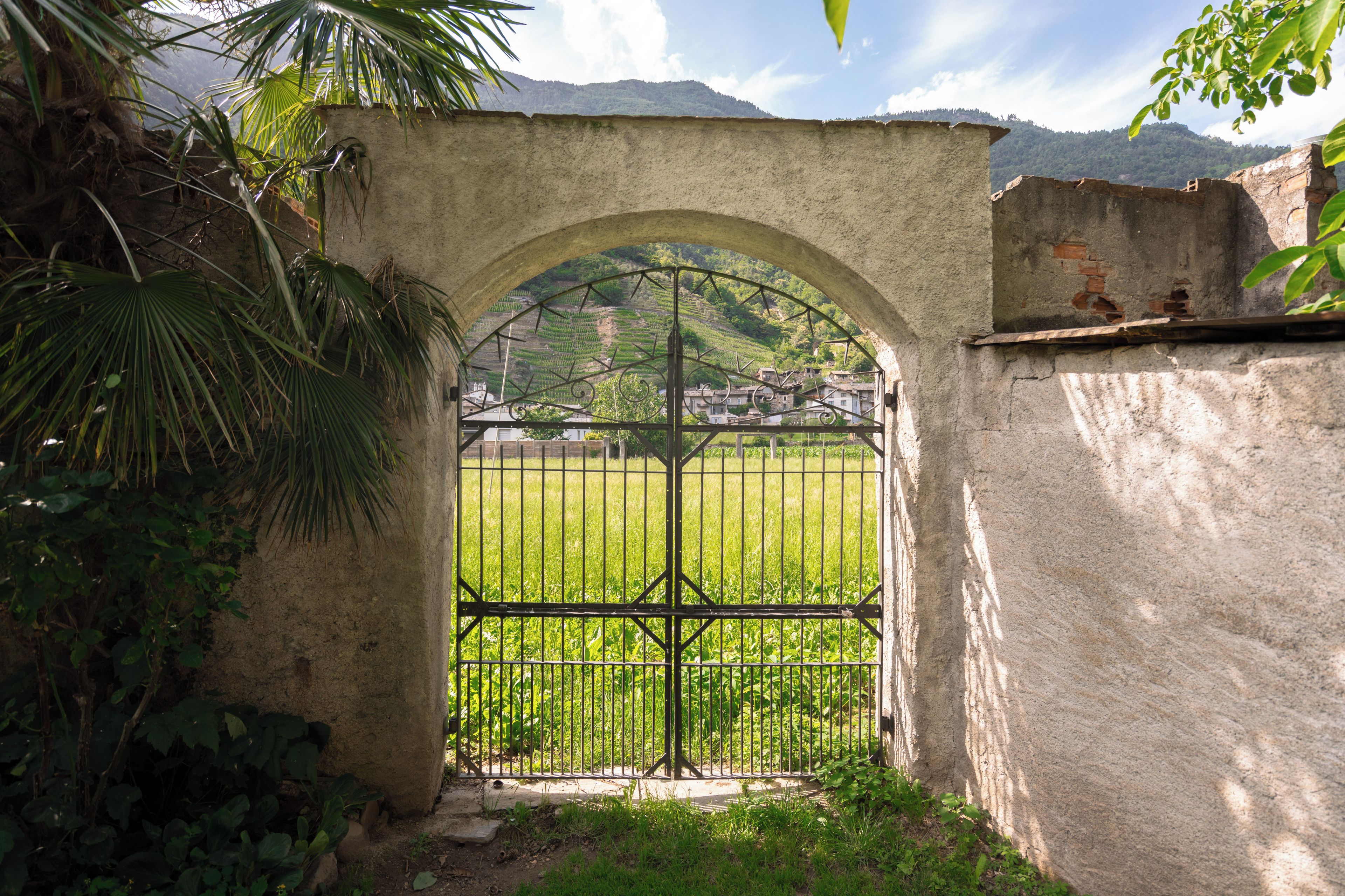 Un cancello rustico con arco e inferriate in metallo, che conduce a una zona erbosa con montagne sullo sfondo.