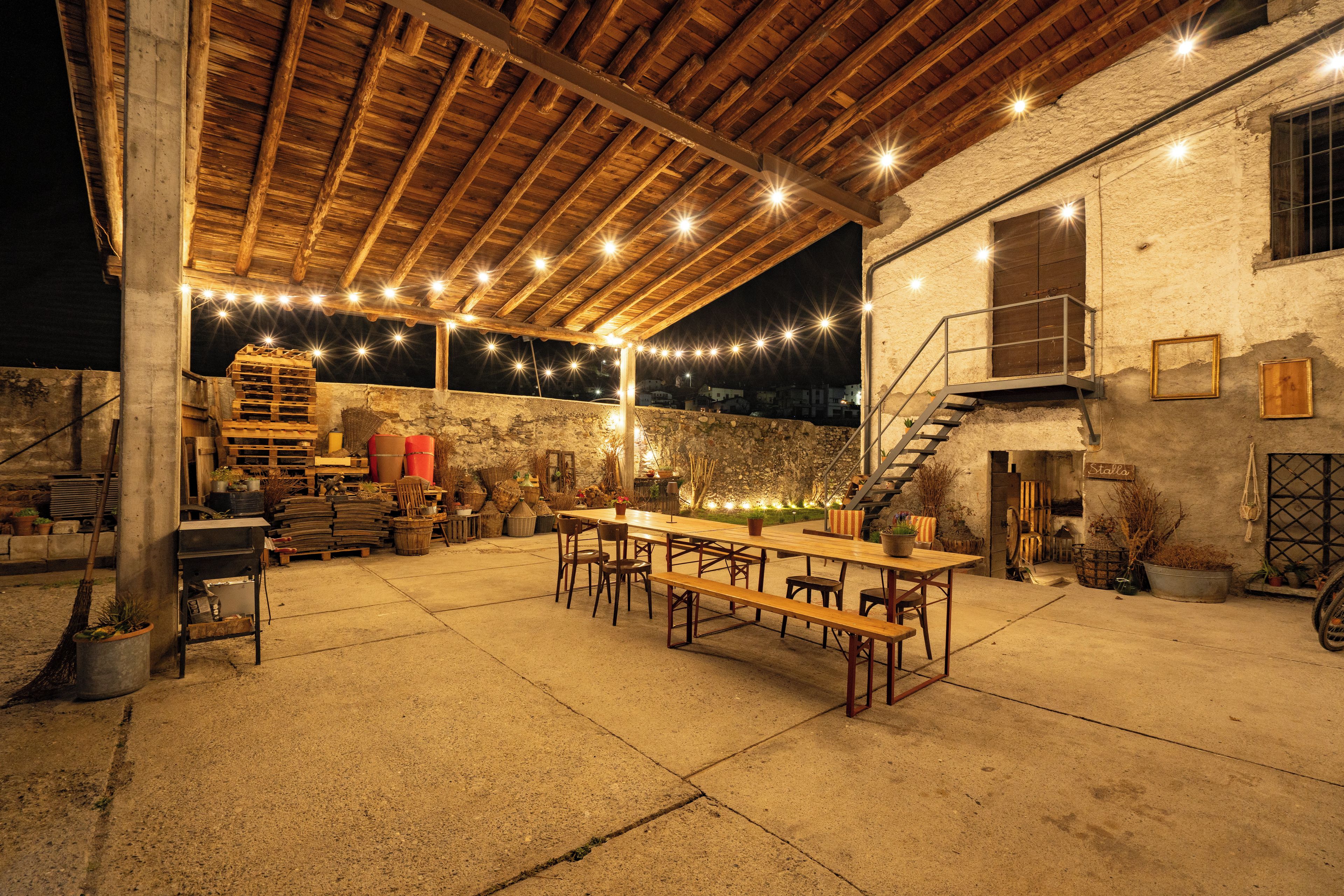 An outdoor patio with a wooden table and benches, surrounded by stone walls and a rustic wooden ceiling.