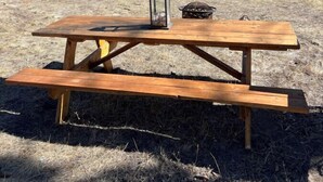 A wooden picnic table with benches, a lantern, and a fire pit in a grassy field with a mountain backdrop.