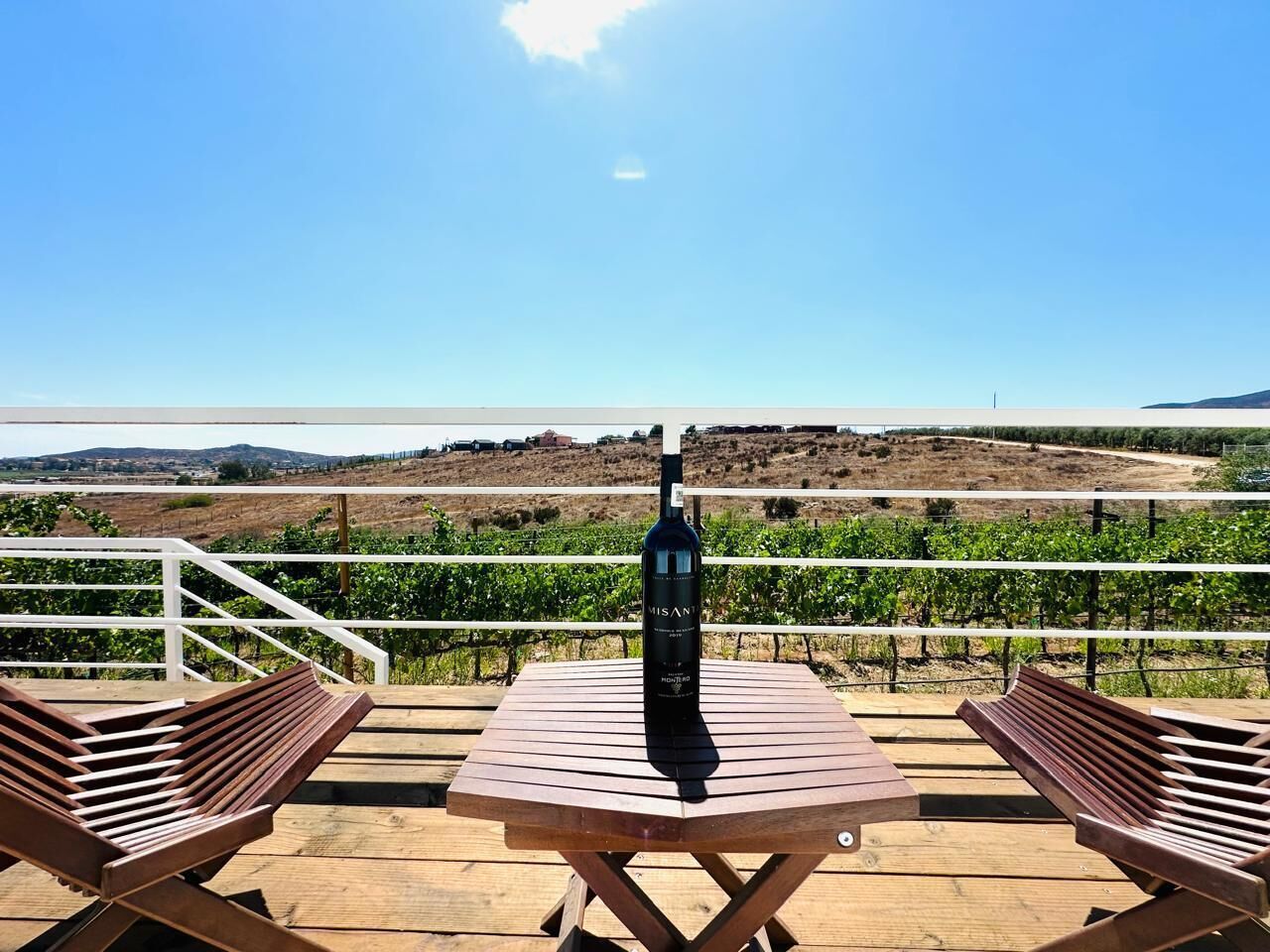 Eine Holzterrasse mit zwei Bänken und einem Tisch, einer Weinflasche und Blick auf einen Weinberg.