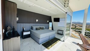 A modern bedroom with a large bed, a nightstand, a desk, and a view of the countryside through a sliding glass door. - Hacienda Montero (Valle de Guadalupe)
