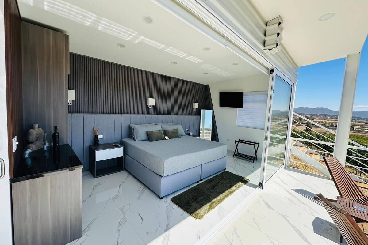 A modern bedroom with a large bed, a nightstand, a desk, and a view of the countryside through a sliding glass door.
