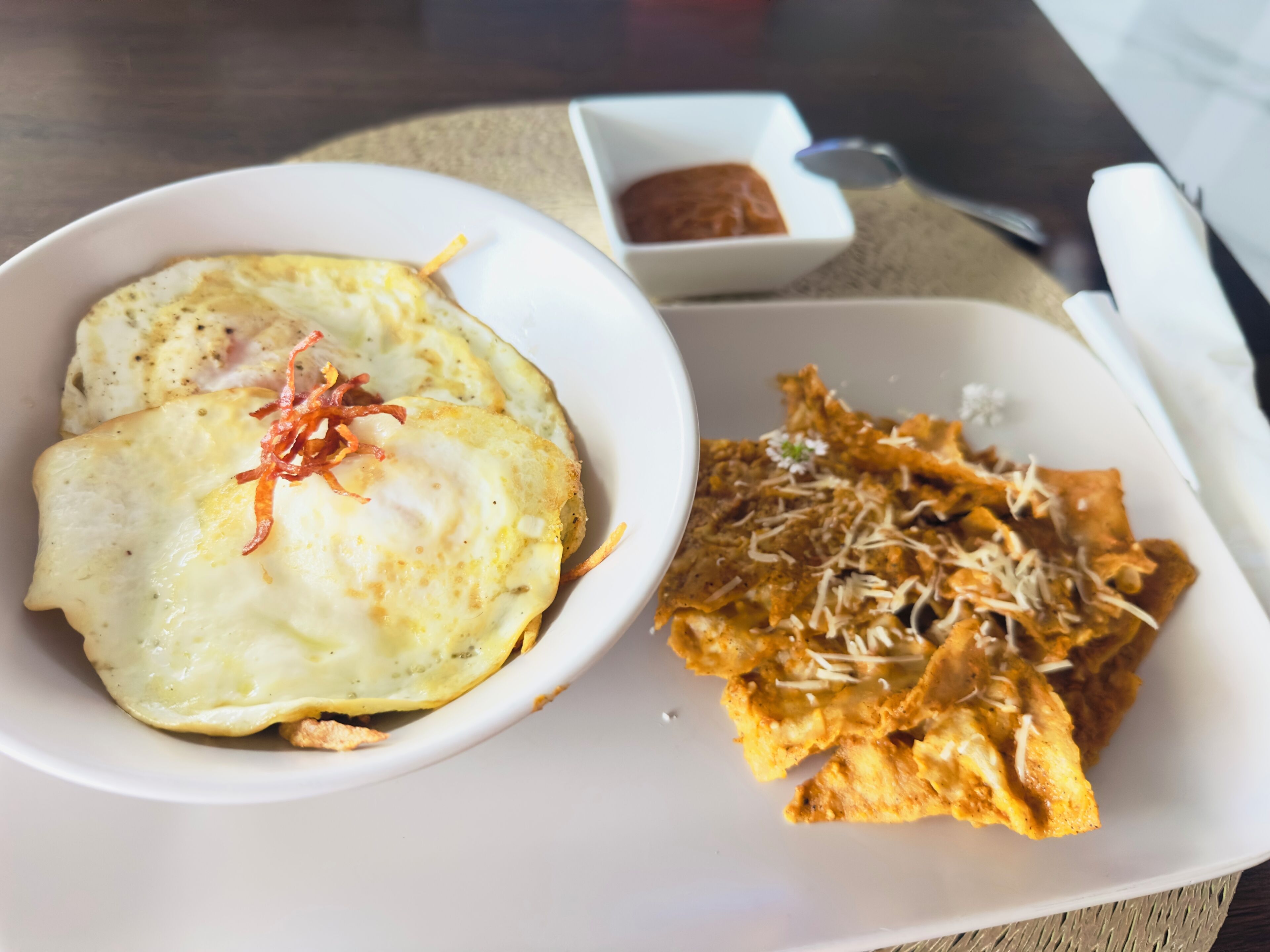 Un plato con dos huevos fritos y una porción de fideos crujientes, decorado con queso rallado y hojuelas de chile rojo.