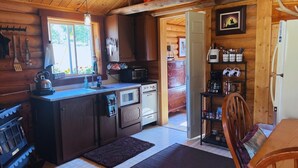 Ein gemütliches Blockhaus-Interieur mit Holzvertäfelung, einer Küchenzeile mit Herd, Spüle und Mikrowelle sowie einem Essbereich mit Tisch und Stühlen.