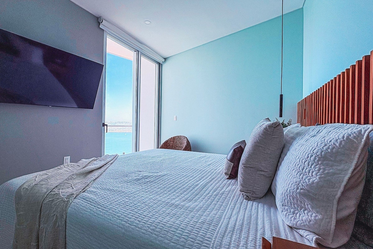 Un dormitorio con cama, televisor en la pared, ventana y vista al mar.