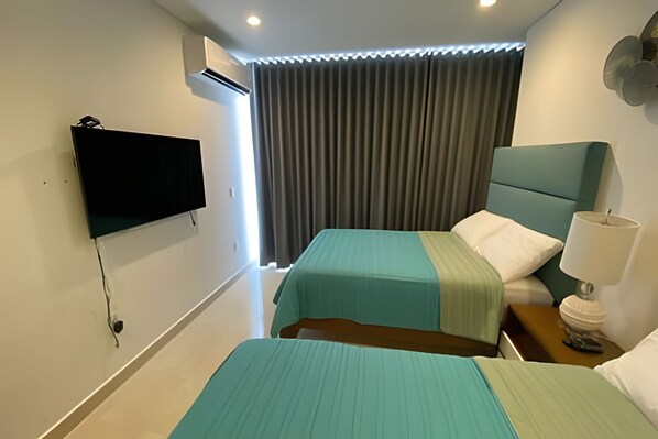 Une chambre d’hôtel avec deux lits, un téléviseur fixé au mur, une fenêtre avec des rideaux et une table de chevet avec une lampe.