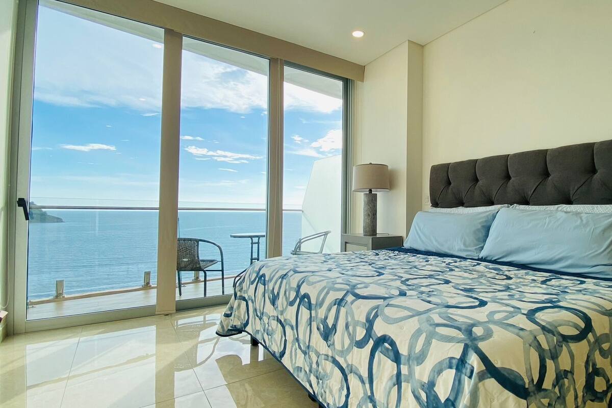 Un dormitorio con una cama grande, una mesita de noche, una lámpara y vista al mar a través de una puerta corrediza de vidrio.