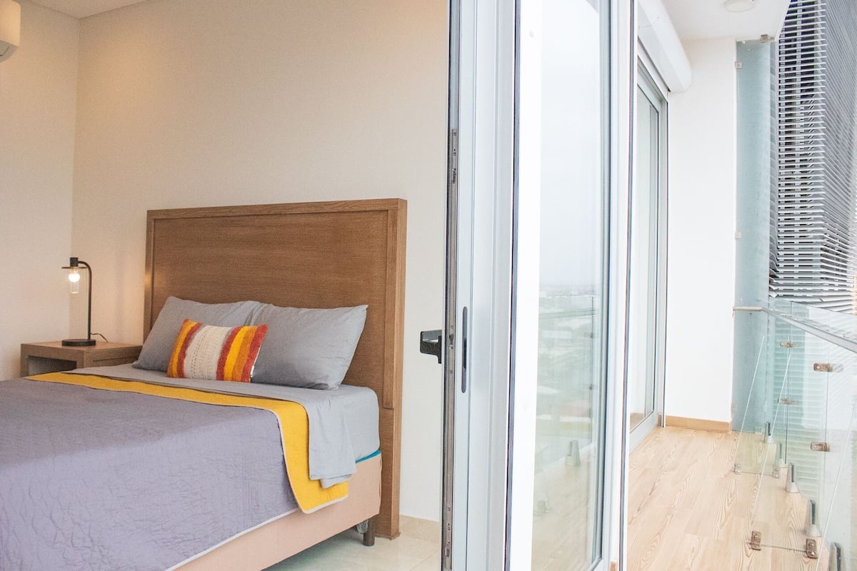 Un dormitorio moderno con cabecera de madera, cama con colcha gris y amarilla, lámpara de noche y un ventanal con persianas.