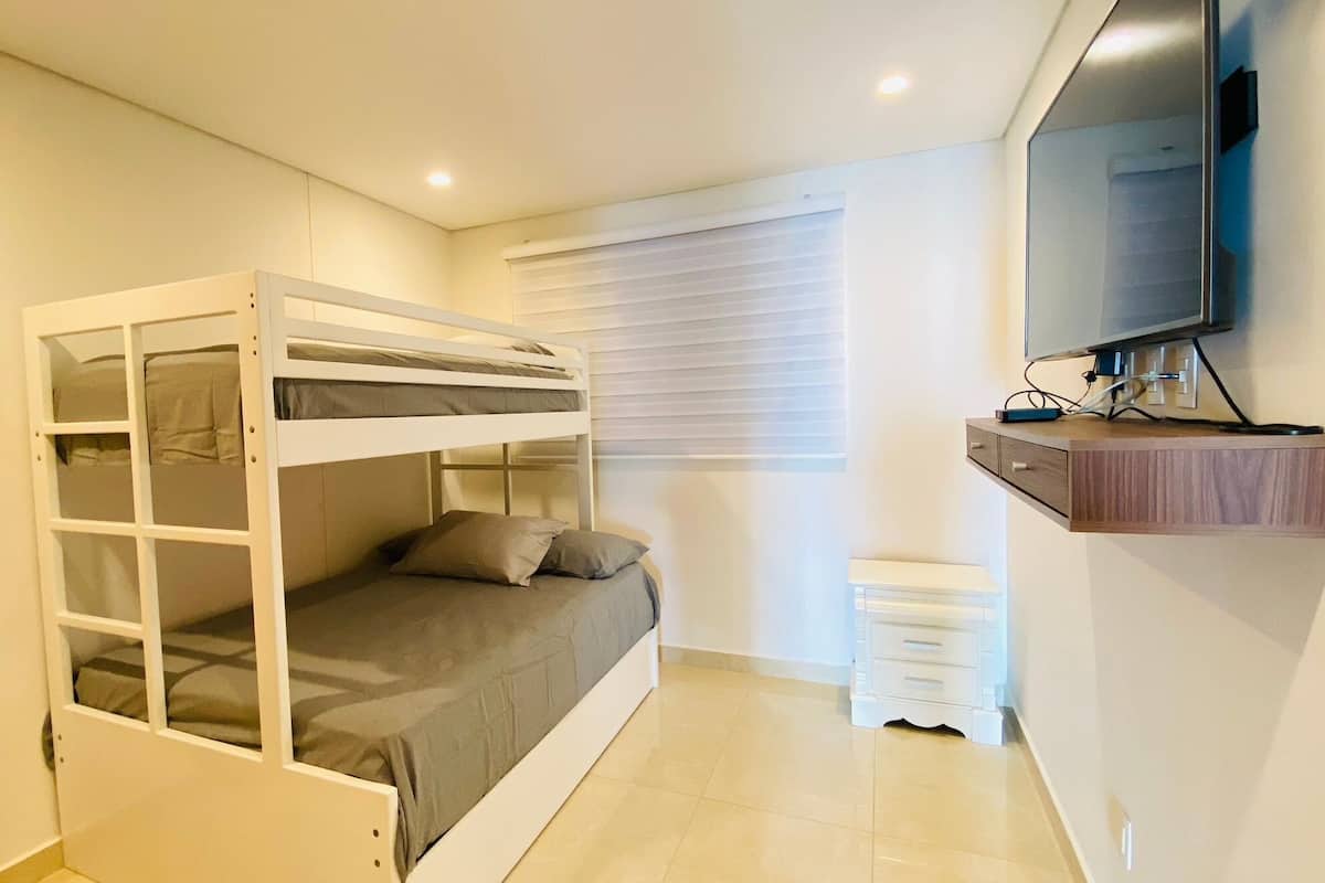 Habitación con literas, un televisor sobre una repisa de madera, una ventana con persianas y un mueble cajonero blanco.