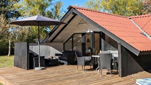 A modern outdoor seating area with a wooden deck, a large umbrella, and a glass-enclosed room with a red roof.