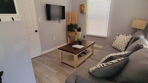 A living room with a gray sofa, a wooden coffee table, a ceiling fan, a TV mounted on the wall, and a window with blinds. - Cozy apartment in charming Denton walkable to TWU (Denton)