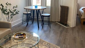 A living room with a round glass coffee table, a beige sofa, a dining table with four chairs, a ceiling fan, and a large window with curtains. - The George 316 an Art Deco Escape (Klamath Falls)