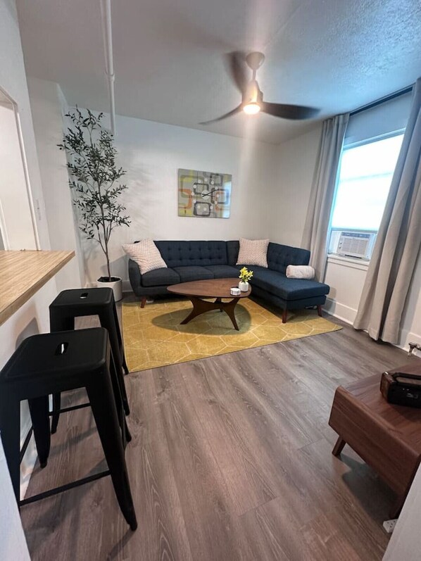 Una sala de estar moderna con un sofá azul oscuro, una mesa de centro de madera, taburetes negros, una planta pequeña y un ventilador de techo.