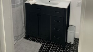 Une salle de bain avec un lavabo blanc, des meubles noirs, un miroir avec éclairage intégré, un rideau de douche et une poubelle.