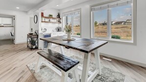 A modern kitchen with white cabinetry, a wooden dining table, and large windows offering a view of the countryside.