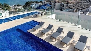 Ein Pool auf dem Dach mit Liegestühlen und Sonnenschirmen mit Blick auf eine Küstenlandschaft, Gebäude und das Meer.